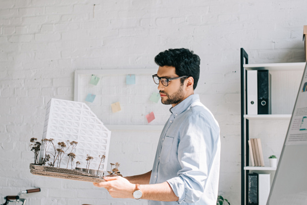 side view of handsome architect in glasses holding architecture model in office - Фото, изображение