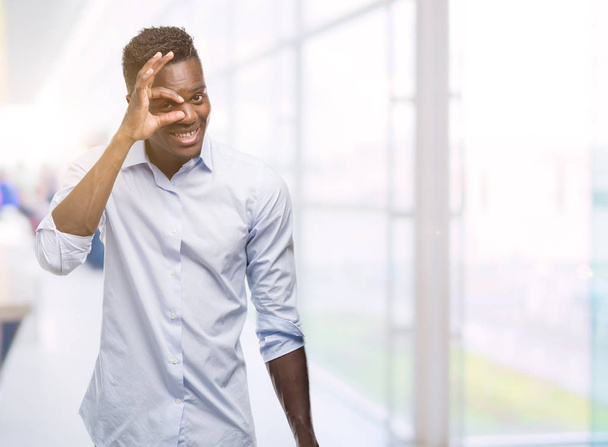 Jonge Afro-Amerikaanse man, het dragen van een shirt met blij gezicht lachend doen ok teken met de hand op oog kijken door de vingers - Foto, afbeelding