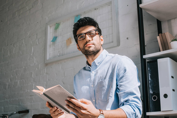 handsome architect in glasses holding book and looking away in office - Fotó, kép