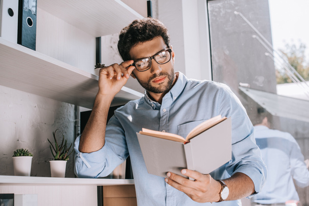 handsome architect touching glasses and reading book in office - Foto, Imagem