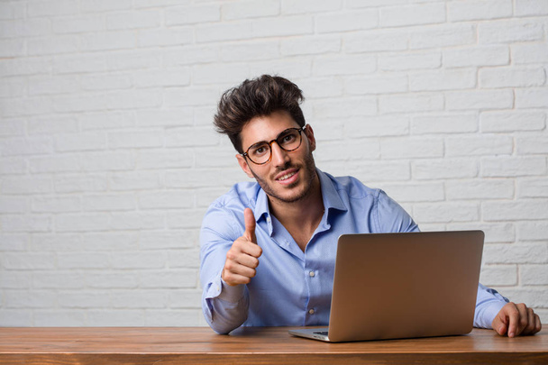 Young business man sitting and working on a laptop cheerful and excited, smiling and raising her thumb up, concept of success and approval, ok gesture - Photo, image