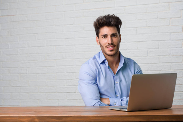 Молодой бизнесмен сидит и работает на ноутбуке, скрещивая руки, улыбаясь и радуясь, будучи уверенным и дружелюбным
 - Фото, изображение