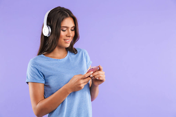 Portrait of a happy young girl holding mobile phone isolated over violet background, listening to music with headphones - Фото, зображення