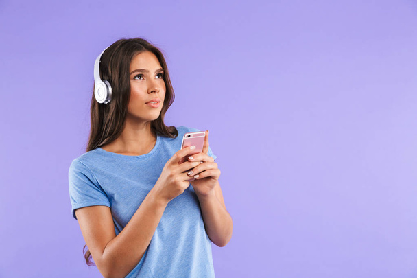 Portrait of a pretty young girl holding mobile phone isolated over violet background, listening to music with headphones - 写真・画像