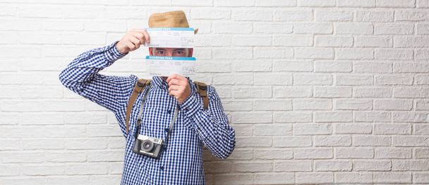 Young traveler man wearing backpack and a vintage camera looking through a gap, hiding and squinting. Holding a boarding pass. - Photo, Image
