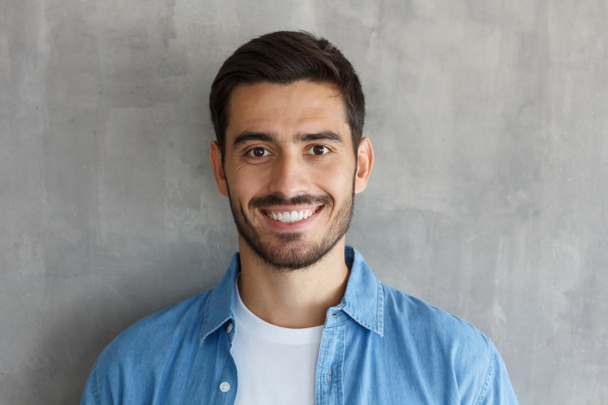 horizontaler Kopfschuss eines jungen europäischen Mannes isoliert auf grauem Hintergrund, der vor der Kamera ein blaues Jeanshemd trägt, freundlich und glücklich lächelnd, zufrieden mit seinem Leben und seinen Freizeitaktivitäten - Foto, Bild