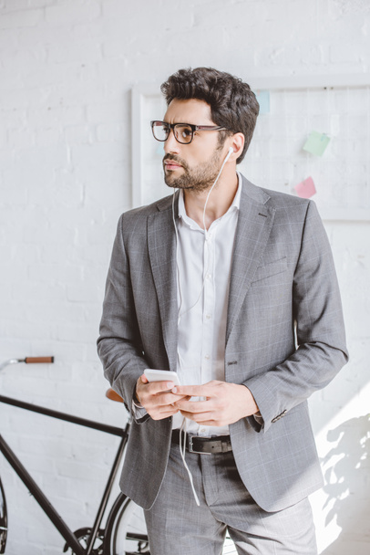 handsome businessman listening music with smartphone and looking away in office - Photo, image