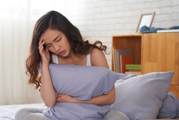 Азиатская женщина сидит на кровати и держит подушку в руке, страдая от головной боли
 - Фото, изображение