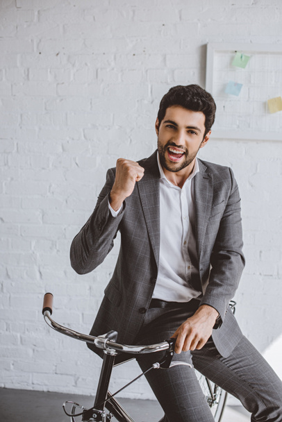 happy handsome businessman sitting on bike and showing yes gesture in office - Foto, immagini