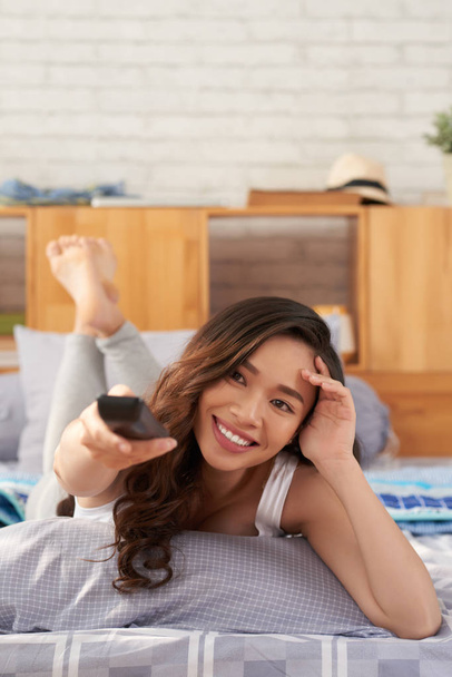 Mulher asiática alegre deitada na cama acolhedora e ligando a TV
  - Foto, Imagem