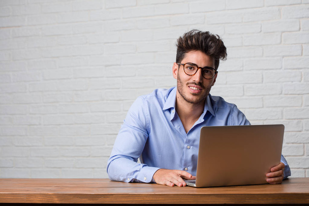 Молодой бизнесмен сидит и работает на ноутбуке весело и с большой улыбкой, уверенно, дружелюбно и искренне, выражая позитивность и успех
 - Фото, изображение