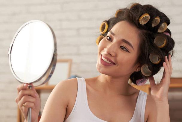 Testa e spalle ritratto di bella donna asiatica che indossa bigodini che guardano allo specchio con ampio sorriso
 - Foto, immagini