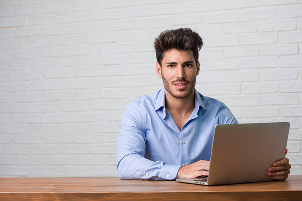 Jonge zakenman zitten en werken op een laptop overschrijding van zijn armen, lachende en gelukkig, zelfverzekerd en vriendelijk - Foto, afbeelding