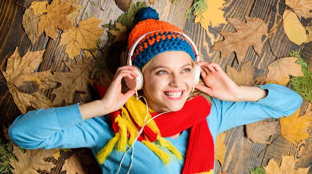 Autumn melody concept. Hipster woman knitted hat and scarf listen music headphones. Fall cozy atmosphere. Fall and autumn season. Girl cheerful face lay on wooden background with leaves top view - Foto, imagen