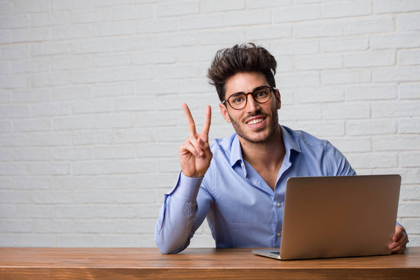 junger Geschäftsmann, der an einem Laptop sitzt und Nummer zwei zeigt, Symbol des Zählens, Konzept der Mathematik, selbstbewusst und fröhlich - Foto, Bild
