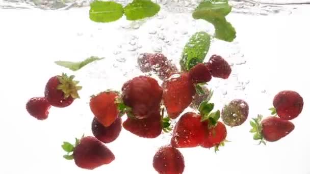 Closeup slow motion video of fresh ripe strawberries and green mint leaves falling and splashing in cold clear water. Concept of freshness - Footage, Video