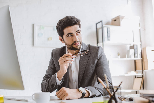 handsome businessman holding glasses and looking away in office - Photo, Image