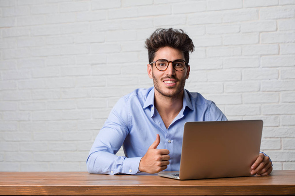 Jonge zakenman zitten en werken op een laptop vrolijk en opgewekt, glimlachend en verhogen haar duim omhoog, concept van succes en goedkeuring, ok gebaar - Foto, afbeelding