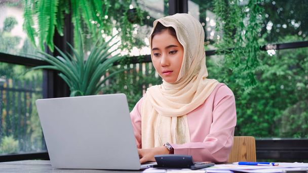 Kaunis nuori hymyilevä aasialainen musliminainen työskentelee kannettavan tietokoneen istuu olohuoneessa kotona. Aasian liiketoiminnan nainen työasiakirja rahoitus ja laskin hänen kotitoimistossa. Kotona viihtyminen
. - Valokuva, kuva
