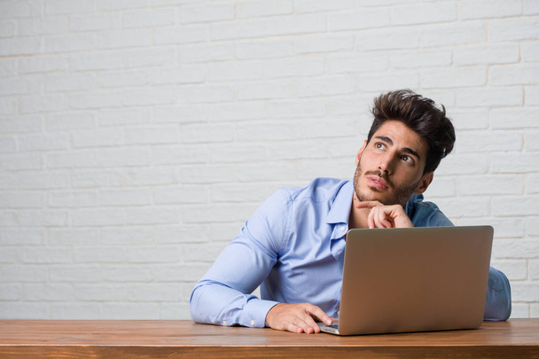 Jeune homme d'affaires assis et travaillant sur un ordinateur portable regardant vers le haut, pensant à quelque chose d'amusant et ayant une idée, concept d'imagination, heureux et excité
 - Photo, image