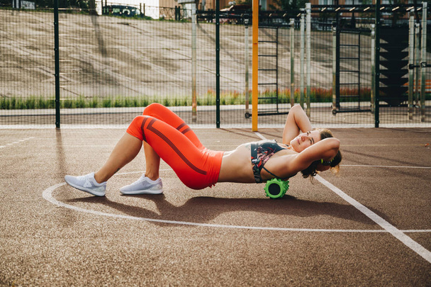 Thema Sport und Reha-Sportmedizin. schöne starke schlanke kaukasische Sportlerin nutzt Schaumstoffrolle Green Field Street Workout, um Schmerzen zu entfernen, zu dehnen und Muskeln zu massieren. - Foto, Bild