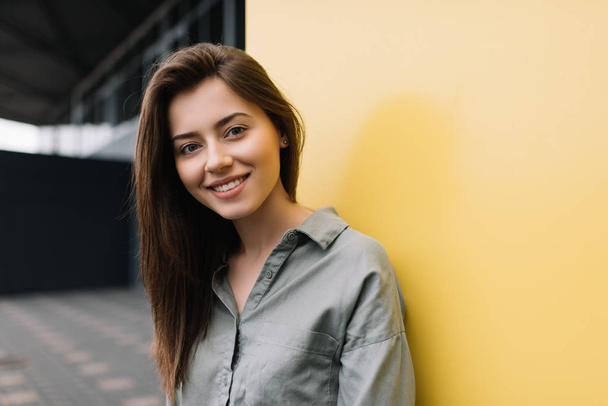 Close up portrait of young happy female wearing casual shirt posing for pictures. Beautiful smiling woman standing on the street near yellow background. Good mood. - Zdjęcie, obraz