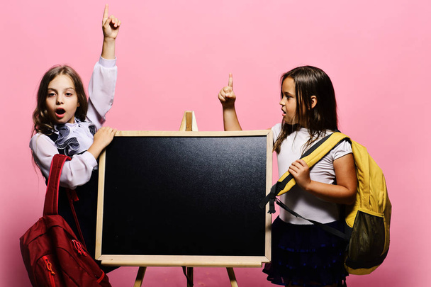 Girls with surprised faces hold fingers up having idea. Kids wearing schoolbags lean on blackboard, copy space. - Фото, изображение
