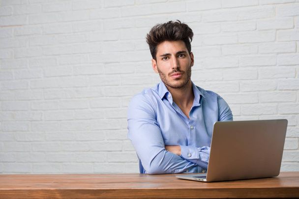 Giovane uomo d'affari seduto e al lavoro su un computer portatile incrociando le braccia, serio e imponente, sentendosi fiducioso e mostrando potenza
 - Foto, immagini
