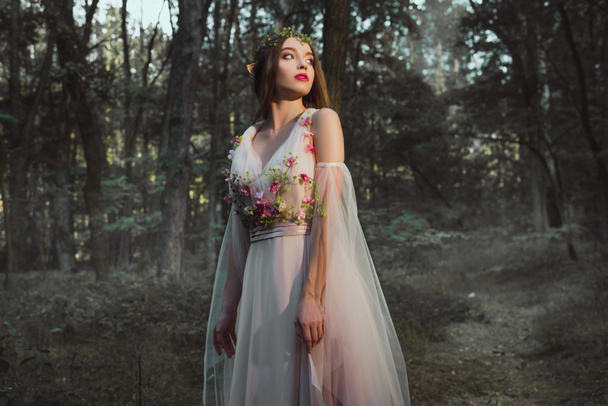 attraktives mystisches Mädchen mit Elfenohren posiert im Blumenkleid im Wald - Foto, Bild