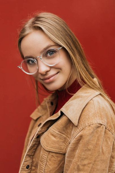 Cheerful positive woman wearing hipster glasses posing for camera near red background. Close up portrait of beautiful female standing on the street - Zdjęcie, obraz