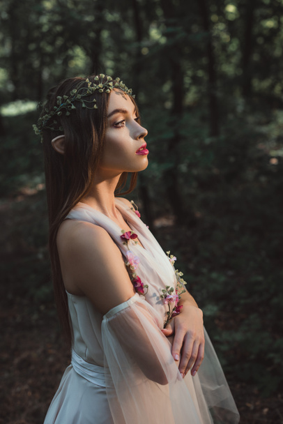attractive mystic elf in flower dress and wreath standing with crossed arms in forest - Photo, Image