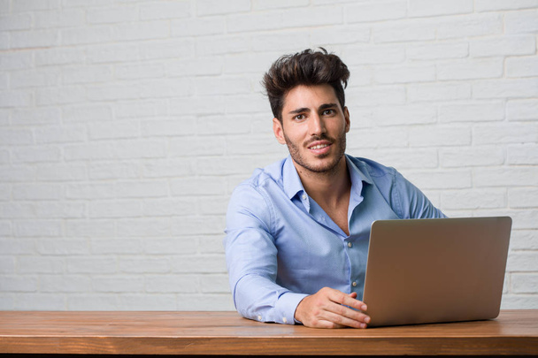 Jonge zakenman zitten en werken op een laptop met de handen op de heupen, permanent, ontspannen en lachende, zeer positief en vrolijk - Foto, afbeelding