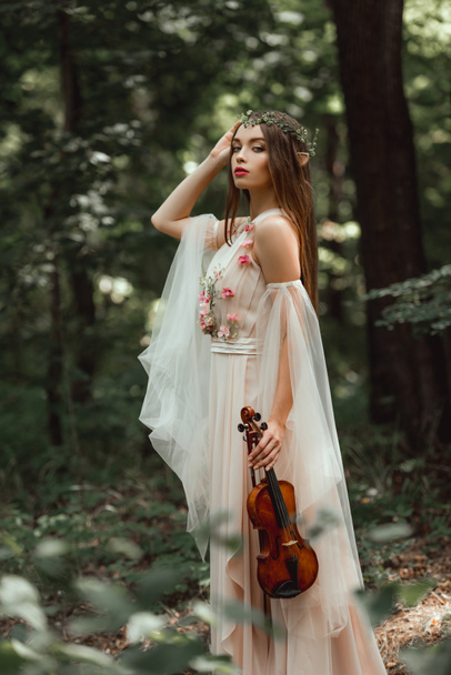 Mystic elf in bloem jurk en bloemen krans holding viool in bos - Foto, afbeelding
