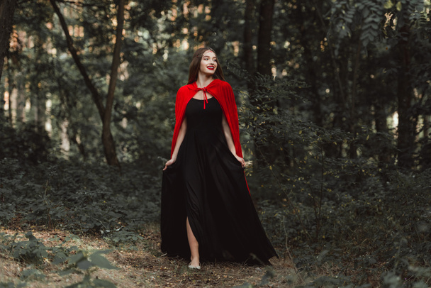 beautiful mystic girl in black dress and red cloak walking in forest - Photo, Image