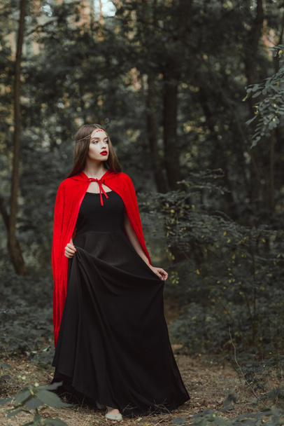 attractive mystic woman in black dress and red cloak walking in woods - Fotoğraf, Görsel