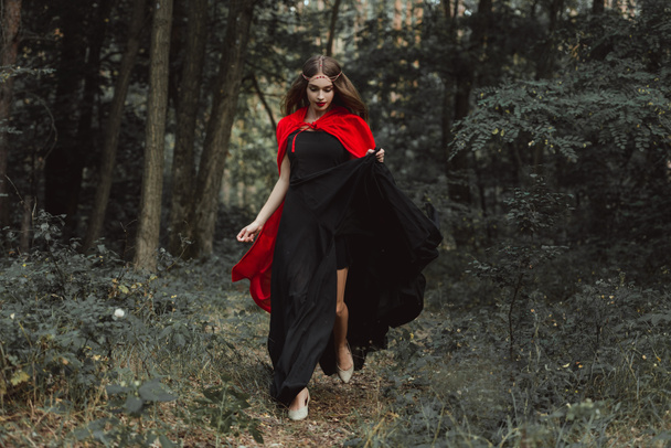 elegant mystic girl in black dress and red cloak running in woods - Photo, Image