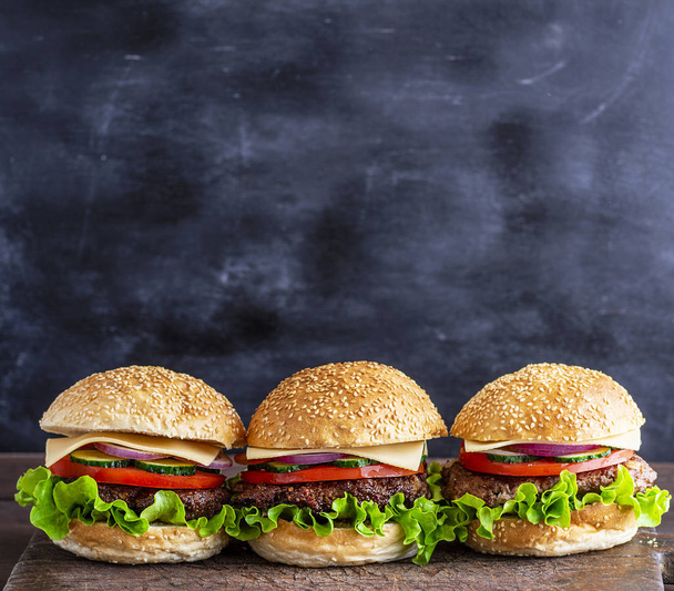 three hamburgers with vegetables on a brown wooden board, behind a black background, empty space - Valokuva, kuva