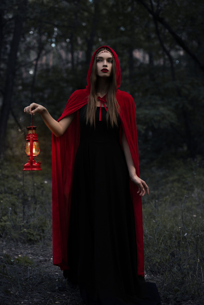 young mystic woman in red cloak with kerosene lamp walking in dark woods  - Fotoğraf, Görsel
