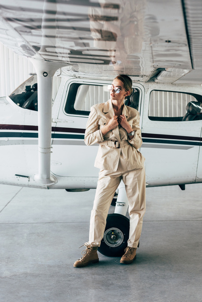 attractive stylish woman in sunglasses and jacket posing near aircraft in hangar - Foto, afbeelding