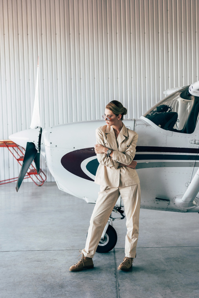 fashionable woman in sunglasses and jacket posing near aircraft in hangar - Foto, imagen