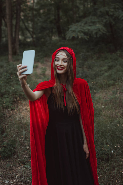 Mystic meisje in de rode mantel selfie nemen op smartphone in bos - Foto, afbeelding