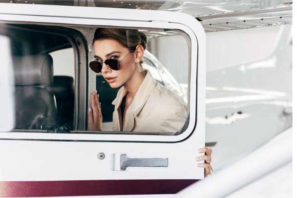 beautiful young woman in sunglasses and jacket looking at camera near aircraft  - Fotó, kép