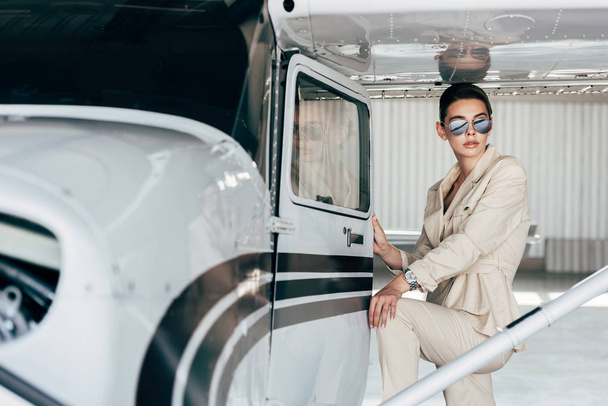 mujer joven con estilo en gafas de sol y chaqueta posando cerca de avión
  - Foto, imagen