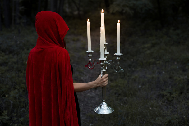 mystic girl in red cloak holding candelabrum with flaming candles in dark woods - Photo, Image