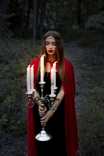 mooi mystic meisje in rode mantel holding kandelaar met brandende kaarsen in bos - Foto, afbeelding