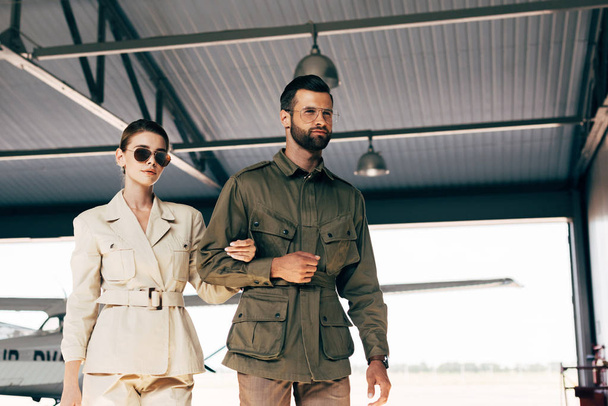 low angle view of fashionable young couple in jackets walking near airplane in hangar  - Photo, Image