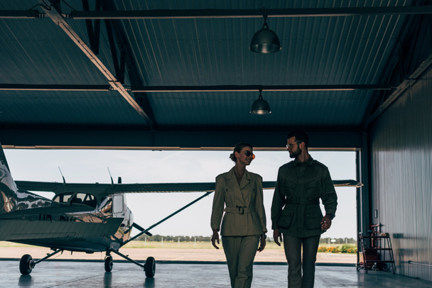 happy stylish young couple in jackets walking near airplane in hangar  - 写真・画像