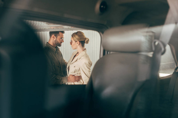 selective focus of young fashionable couple in jacket embracing and looking at each other near plane  - Foto, Imagen