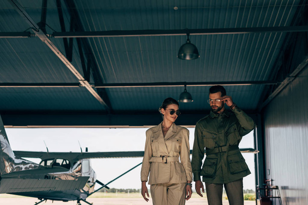 couple in stylish jackets walking near airplane in hangar  - Photo, Image