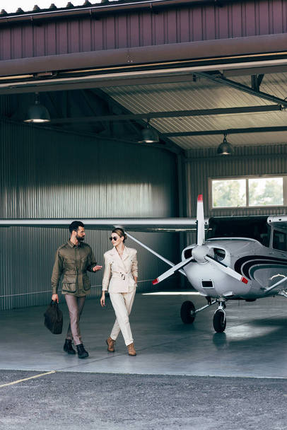 young man with bag gesturing by hand and talking to attractive girlfriend in hangar with plane  - Fotografie, Obrázek